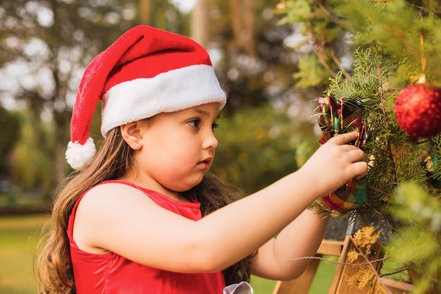 クリスマスの帽子をかぶった愛らしい少女がクリスマスツリーにボールを置きます