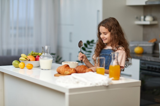 Прелестная маленькая девочка с вьющимися волосами ест здоровый завтрак хлопьев на кухне.