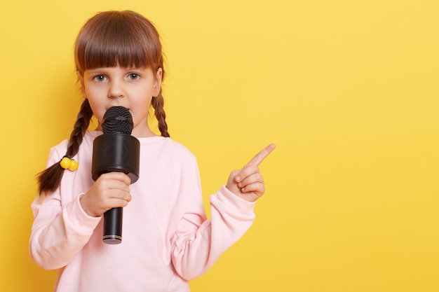 Photo adorable little girl with microphone on yellow wall, while talking in mic, pointing index finger aside. copy pace .