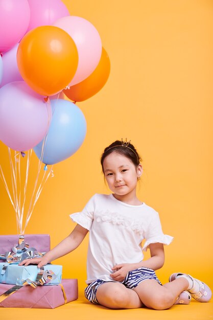 Adorable little girl with small crown on head sitting in isolation by pile of birthday gifts and bunch of balloons