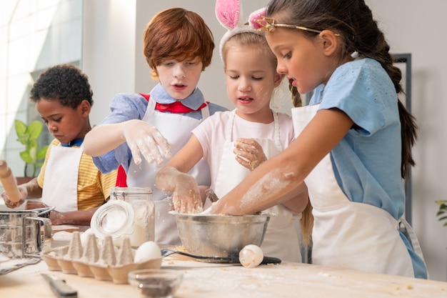 Adorable little sisters looking at camera with wide smiles while standing at wooden table and preparing healthy snack for their parents