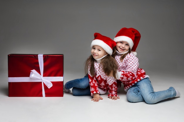 Adorabili sorelline che portano il cappello rosso della santa che si abbracciano sul pavimento con un grande regalo di natale in carta rossa e muro grigio bowon bianco.