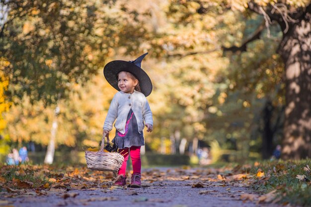Adorable little toddler girl in autumn park girl in witch costume and black hat cosplay halloween