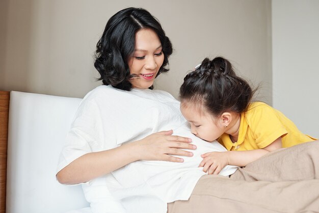 Adorabile bambina vietnamita che bacia la pancia della madre incinta