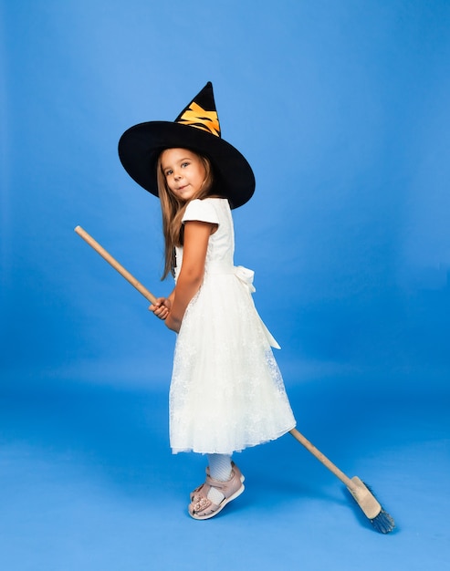 Adorable little witch in a white dress flies on a broomstick isolated on a blue background.