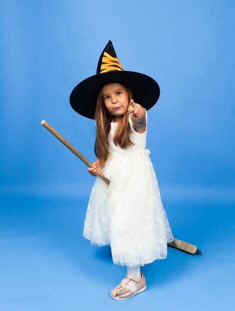 Adorable little witch in a white dress flies on a broomstick isolated on a blue background.