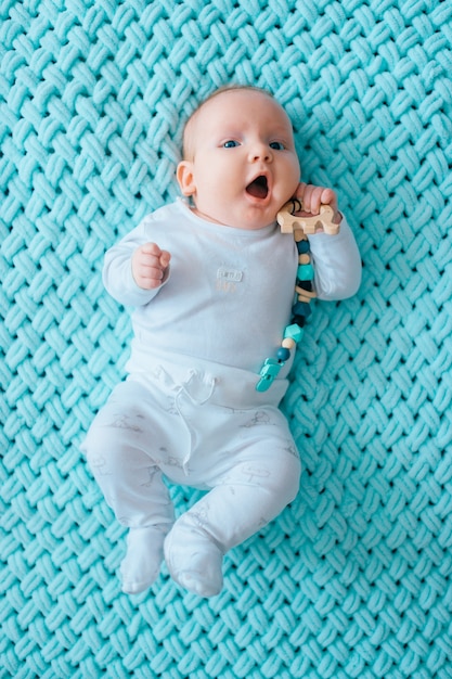 Adorable lovely newborn male baby with blue eyes lifestyle  indoor portrait from above. Cute child with plump cheeks lying on back in white pijama on turqoise woolen plaid. Funny nursing kid emotions.