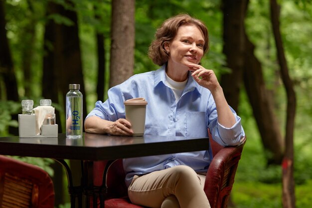 Adorable mature woman sits at the table in outside cafe with paper coffee cup and bottle of water se