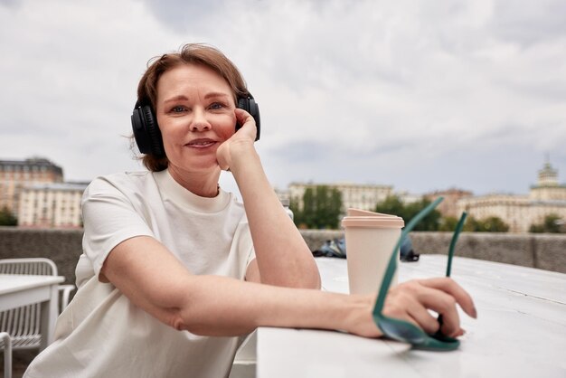 Foto adorabile donna di mezza età in maglietta bianca con occhiali da sole, cuffie wireless e tazza di carta da caffè