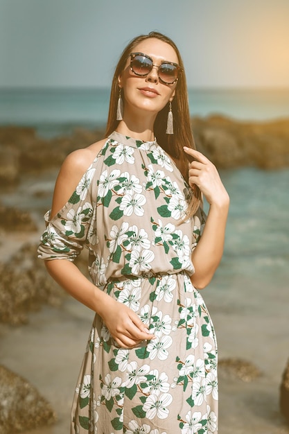 Очаровательная мисс в летнем цветочном сером платье и черных солнцезащитных очках позирует на пляже.