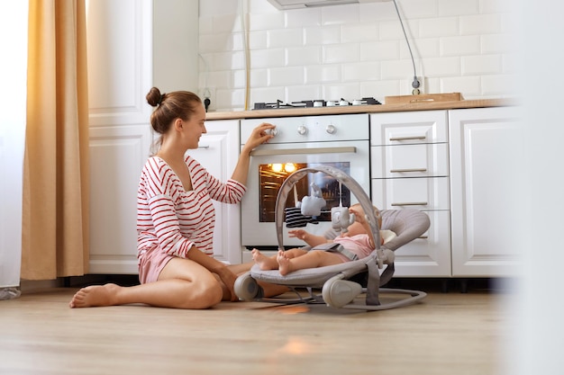 Adorabile madre che indossa una camicia a righe casual e una breve seduta sul pavimento con la figlia del bambino sulla sedia a dondolo, imposta la temperatura nel forno, prepara una torta o una torta.