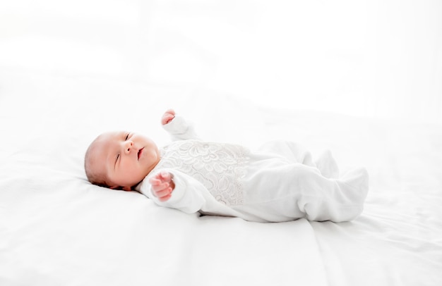 ベッドに横になってカメラを見ている白い衣装を着た愛らしい新生児の女の子。家で休んでいるかわいい幼児の子供