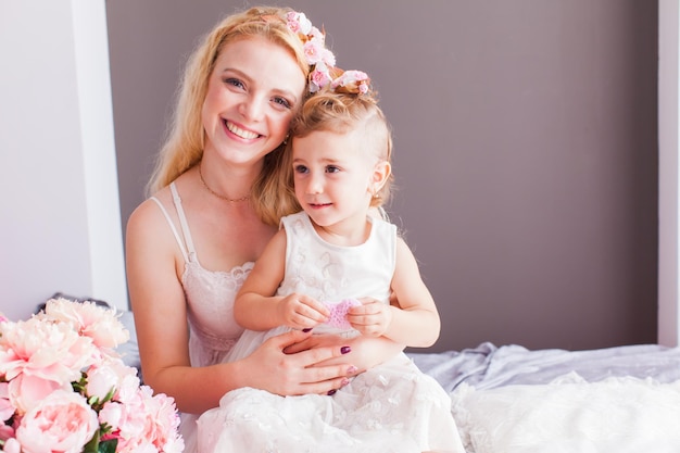 Adorable portrait of young mother and her daughter sitting on the bed in their house. Attractive young mother holding her daughter looking at the camera, both wearing white dress, birthday concept