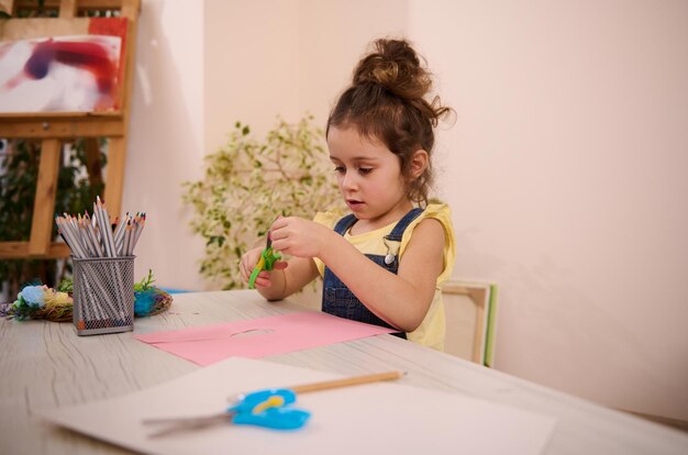 Adorable preschool age European 4 years old girl cuts out shapes from colored paper making appliques during art class at home Kids entertainment education art and creativity concept