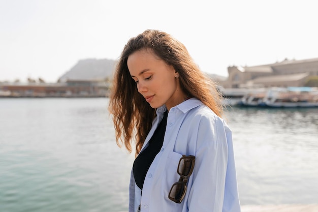 Foto adorabile donna graziosa con acconciatura lunga ondulata volante che indossa una camicia blu sta guardando in basso con un sorriso meraviglioso mentre cammina sul molo alla luce del sole in una calda giornata di sole concetto di riposo e relax