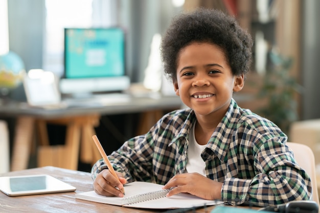 Adorable schoolboy of African ethnicity making notes in copybook and looking at you with smile while doing homework by table in living-room