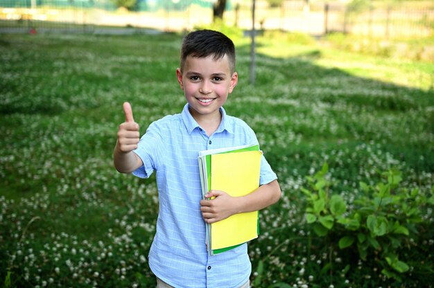 Фото Очаровательный школьник держит желтую тетрадь и показывает большой палец в камеру, стоящую на фоне зеленой травы