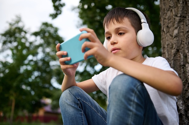 Adorable schoolboy wearing wireless headphones, playing with mobile phone, resting on the green grass of urban park