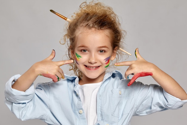 Adorable schoolgirl having a brush in her chic curly blond hair, wearing in a blue shirt and white t-shirt. She has painted her cheeks and pointing on them, smiling at the camera on a gray background.