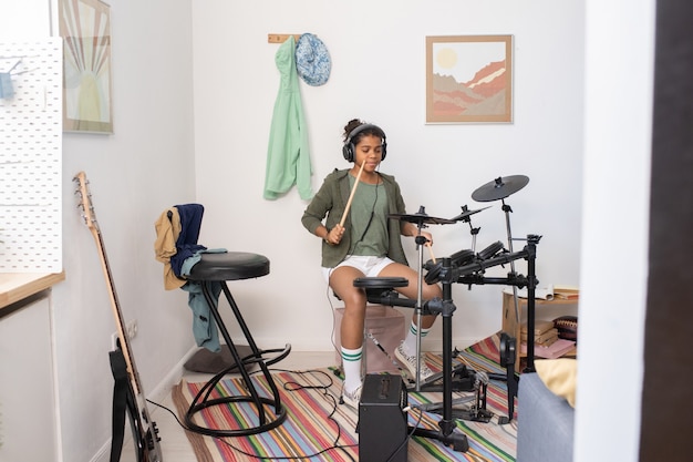 Adorable schoolgirl in headphones and casualwear playing drumkit while having lesson of music at home or studio