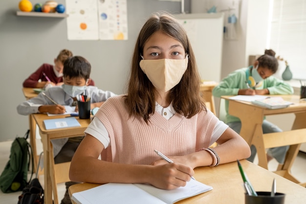 Adorable schoolgirl looking at you while making notes at lesson