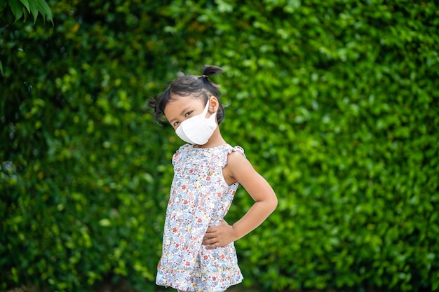 Adorable small girl with healthy face mask standing against green nature background