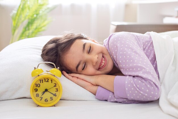 A adorable smiling brunette girl in purple polka dot pajamas lie on a white pillow on a bed