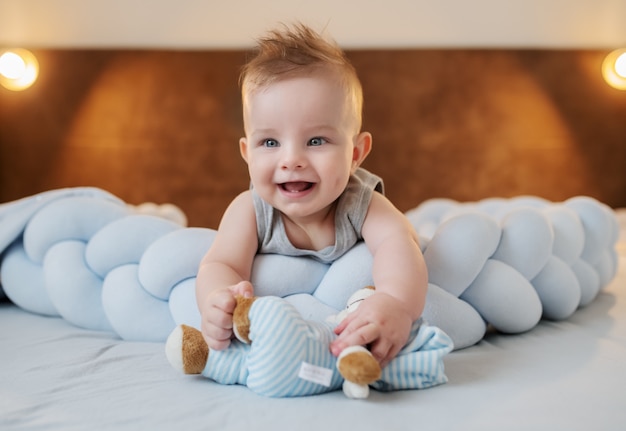 Adorable smiling caucasian little baby boy 6 months old lying on stomach on bed in bedroom and holding teddy bear.