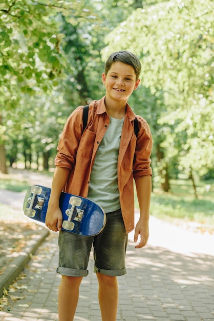 Photo adorable smiling schoolboy holding skateboard and looking at camera in park
