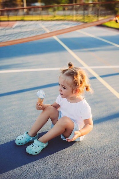 스포츠 코트에서 야외에서 아이스크림을 먹는 사랑스러운 세련된 유아 소녀