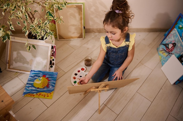 Adorable sweet baby girl drawing picture on canvas sitting on the floor around beautiful child painted images Art class creativity motor skills development creativity and kids education concept