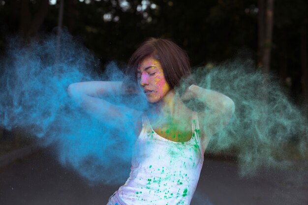 Adorable tanned woman with short hair posing with exploding Holi blue and green dry paint