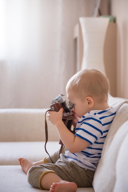 Adorable toddler boy sitting on the sofa in the living room and playing with vintage photo camera