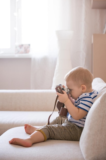 Adorable toddler boy sitting on the sofa in the living room and playing with vintage photo camera