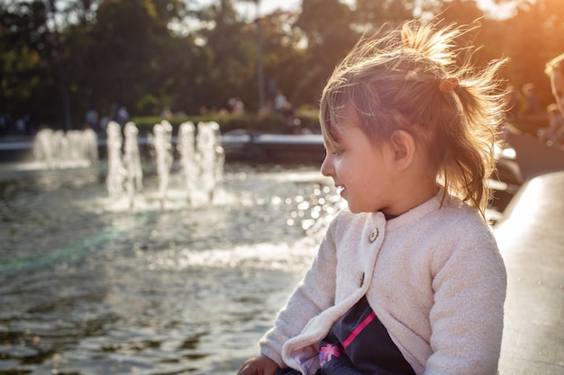 愛らしい幼児の女の子は、晴れた日に公園の噴水のある池を見ます。週末の家族の散歩。子供たちと過ごす時間。芸術的な焦点