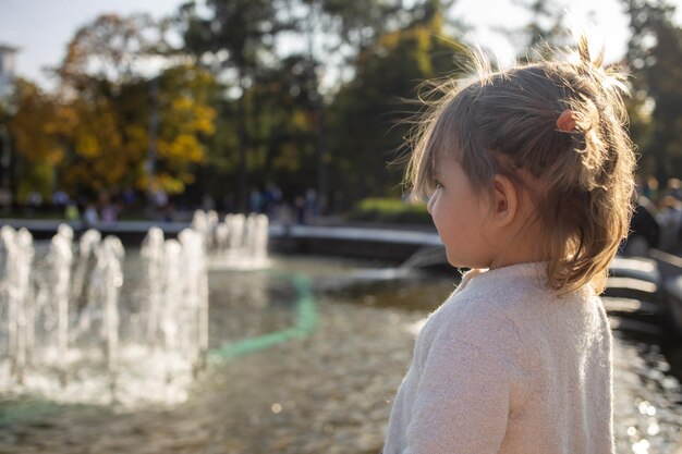 愛らしい幼児の女の子は、晴れた日の週末の家族の公園で噴水のある池を見ます