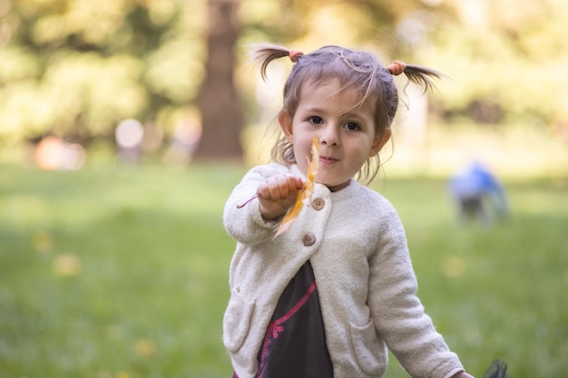 Adorable toddler girl smiles at the camera and holds out a yellow autumn leaf in an autumn park