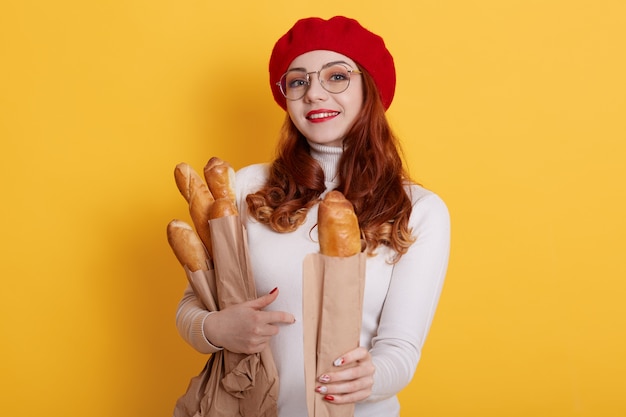 Adorable woman holding paper bag with long bread baguettes, offers one to somebody, wearing white shirt, red beret and glasses on yellow