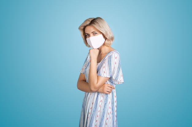 Adorable woman with blonde hair wearing a white medical mask is posing at camera on a blue wall at s...