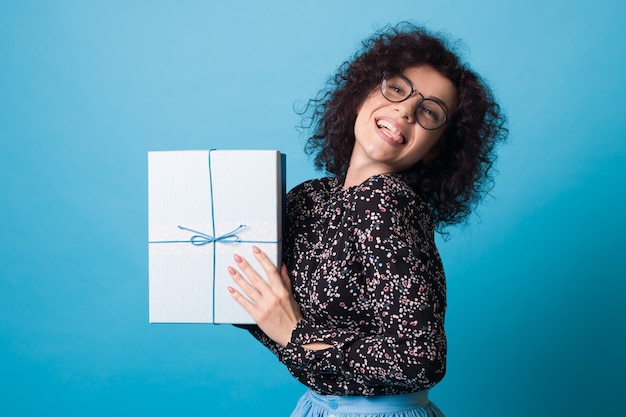 Photo adorable woman with curly hair and glasses is showing her tongue while holding a present on a blue  wall