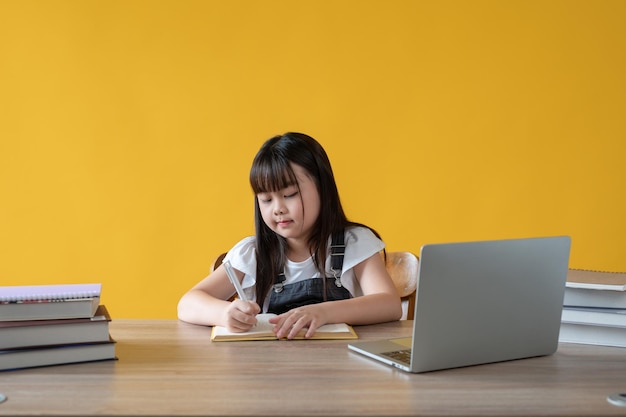 An adorable young asian girl is doing homework at her study table isolated yellow background