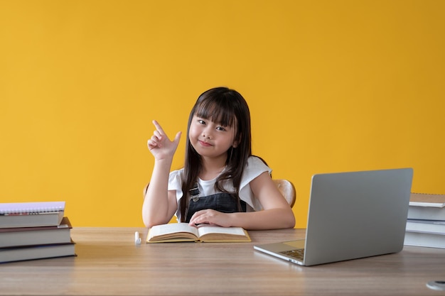 An adorable young asian girl sits at her study table with schoolbooks and a laptop on the table