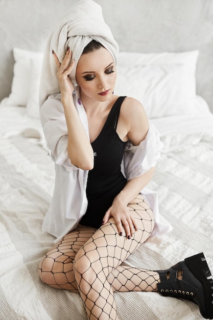 Adorable young caucasian woman with bright makeup and a towel on her head, wearing bodysuits, mesh pantyhose and trendy boots posing on the bed in the morning