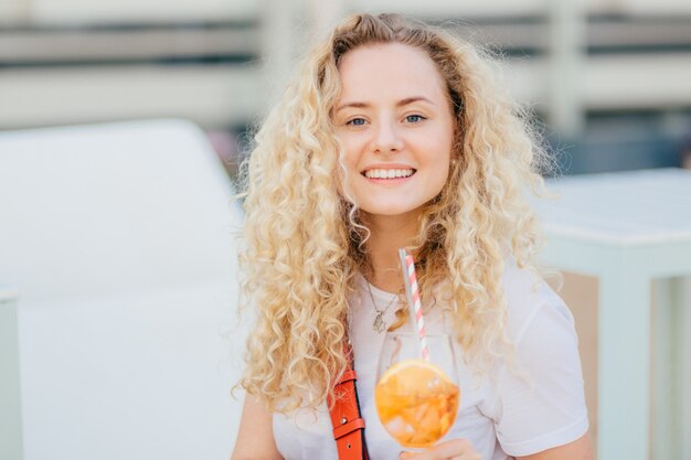 Adorable young female has toothy smile, fresh skin, blue eyes, drinks summer fresh cocktail, looks happily at camera, has unforgettable rest with friends during summer vacation. Leisure concept