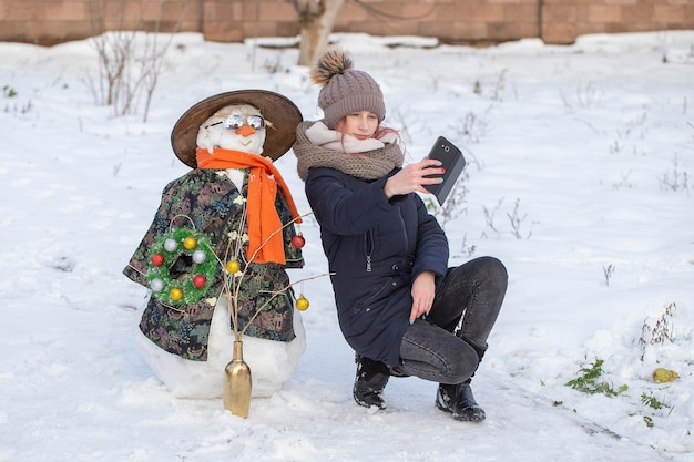 Adorable young girl is taking pictures of selfie with a snowman in beautiful winter park Winter activities for children Closeup