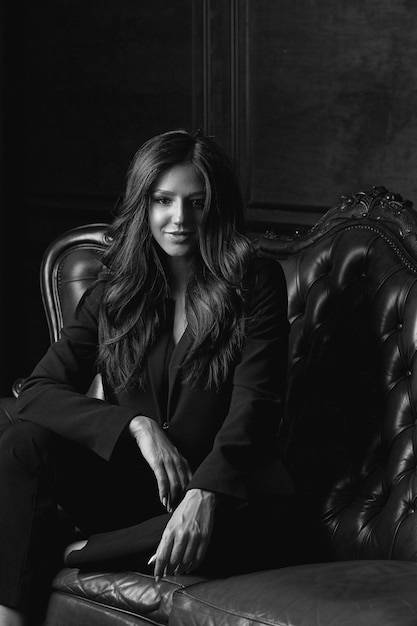 Adorable young model with long hair wearing jacket posing on a vintage sofa. Black and white toning