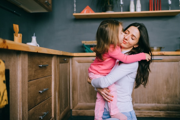 Adorable young mother hugs her little funny daughter in kitchen. Happy family indoor lifestyle portrait.