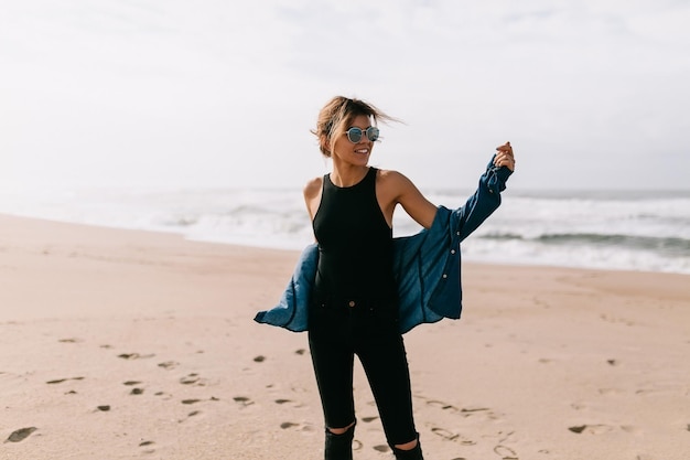 Adorable young stylish European woman in black outfit in denim shirt is posing on the sandy shore near the ocean on clear blue sky background