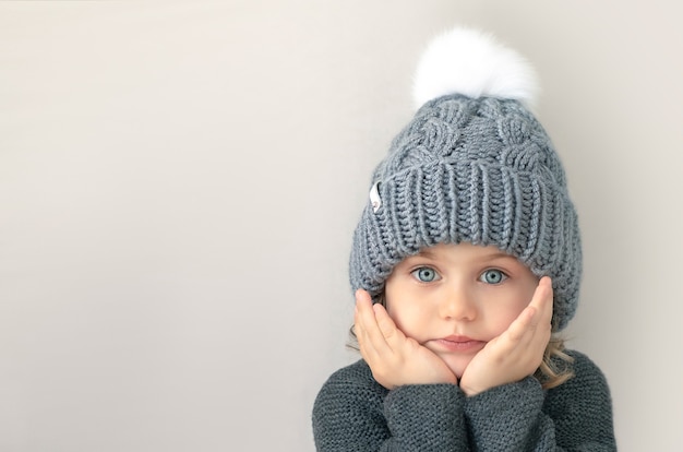 Adorible child girl with big blue eyes in grey wool knitted sweater and hat with white pompom looks at camera with hands on cheeks