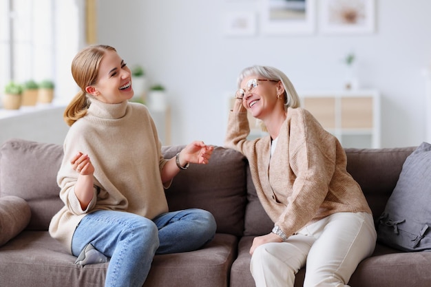 Adult and aged women laughing out loud at joke while relaxing on comfortable couch in cozy living room at home